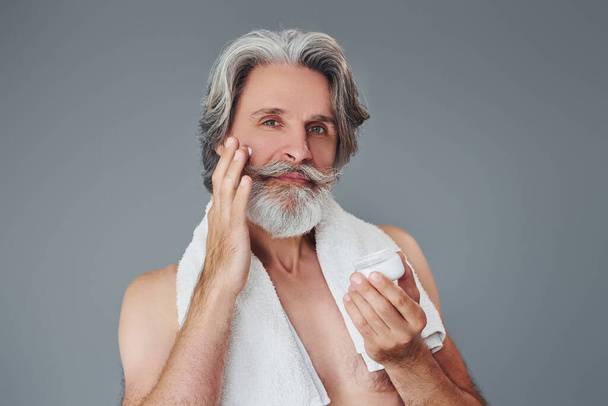Taking care of face and skin clearness. Stylish modern senior man with gray hair and beard is indoors. - Фото, изображение