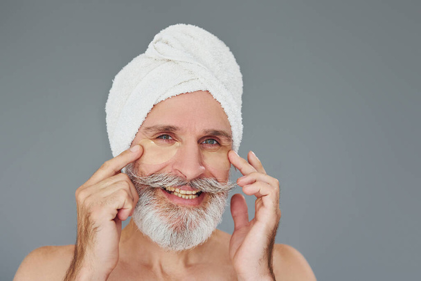 With white towel on head. Stylish modern senior man with gray hair and beard is indoors. - Foto, imagen