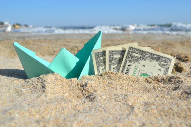 Barco de papel pequeno azul e notas de papel de três dólares metade na areia na praia near sea - Foto, Imagem