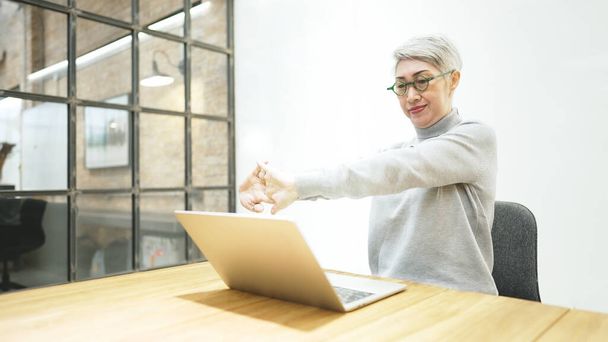 Концепція офісного синдрому. Зріла азіатська бізнес-леді носить окуляри, використовуючи ноутбук, сидячи за робочим столом. Вона розтягує руку і розслабляє шию
 - Фото, зображення