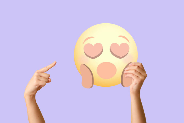 Manos femeninas sosteniendo emoticono sorprendido con ojos en forma de corazón sobre fondo lila - Foto, Imagen