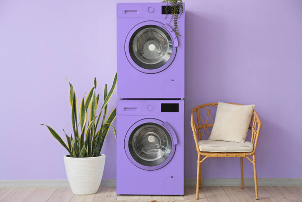 Інтер'єр сучасної пральної кімнати з двома пральними машинами, стільцем і кімнатами біля бузкової стіни
 - Фото, зображення