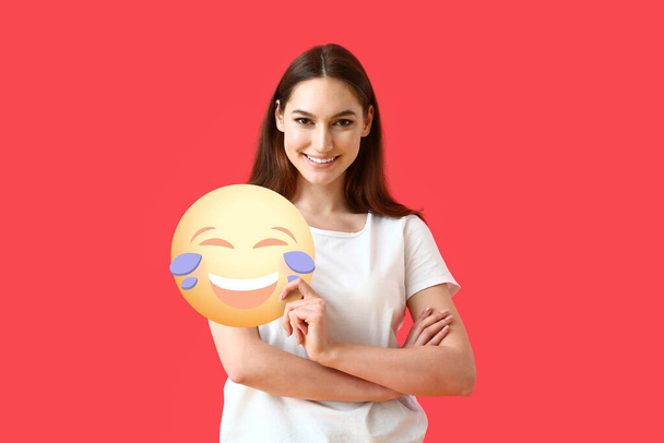 Hübsche junge Frau hält Emoticon mit Freudentränen auf rotem Hintergrund - Foto, Bild