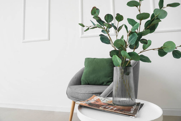 Table with eucalyptus in vase, magazine and armchair near light wall - Foto, imagen
