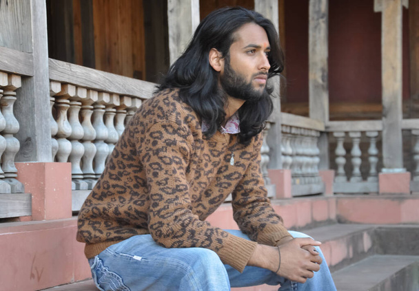 Side view of a good looking long haired Indian young guy, looking sideways while sitting in temple stairs with leaning forward - Foto, imagen