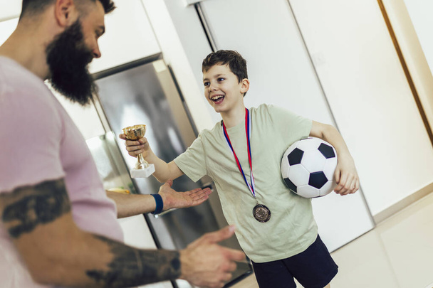誇り高い父はサッカートーナメントの後に彼の息子のメダルとトロフィーを見てとても興奮しています. - 写真・画像