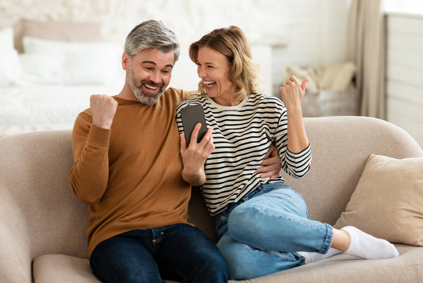 Щаслива середнього віку пара використовує смартфон трясе кулаками вдома
 - Фото, зображення