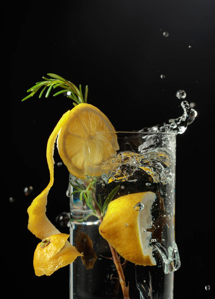 Τζιν τονωτικό κοκτέιλ με λεμόνι και δενδρολίβανο. Φέτα λεμόνι πέφτει σε ποτήρι με ένα κοκτέιλ. - Φωτογραφία, εικόνα