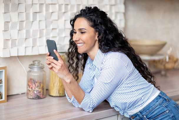 Счастливая латинская женщина в случайном использовании смартфона, серфинг в социальных сетях, общение с друзьями, проводить время на кухне - Фото, изображение