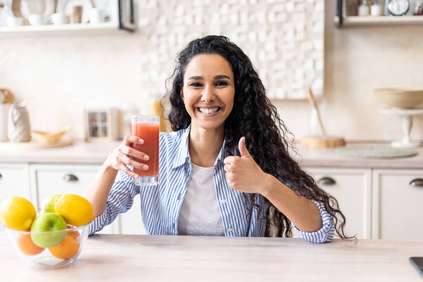 Mulher latina positiva segurando vidro de suco de tomate orgânico e mostrando o polegar para cima, sentado à mesa na cozinha leve - Foto, Imagem