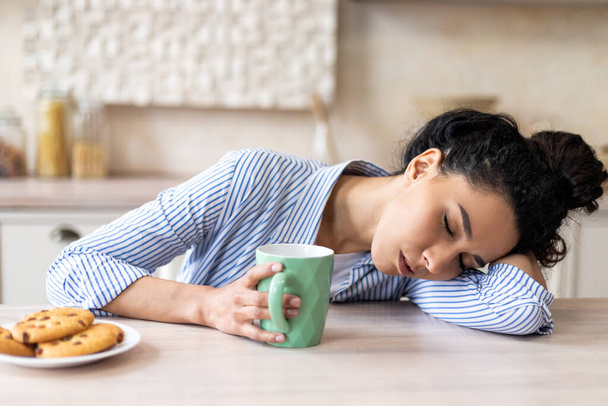 Femme latine fatiguée avec tasse à café, se réveillant tôt et dormant sur la table dans la cuisine intérieure légère, espace libre - Photo, image