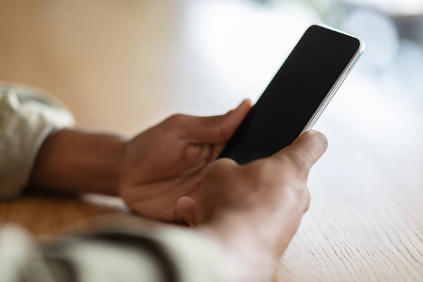 Millennial hombre negro escribiendo en el teléfono inteligente con pantalla en blanco en la mesa de madera en el interior de la cafetería, irreconocible - Foto, imagen