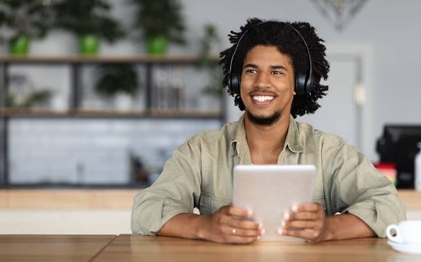 Vrolijke glimlachende krullende jonge zwarte man in draadloze hoofdtelefoon op tablet, muziek luisteren en kijken naar lege ruimte - Foto, afbeelding