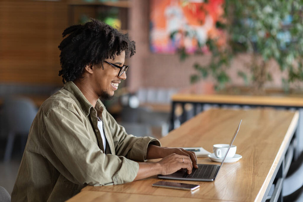 Vrolijke glimlachende krullende millennial zwarte man in glazen typen op laptop aan tafel in cafe interieur, kopiëren ruimte - Foto, afbeelding