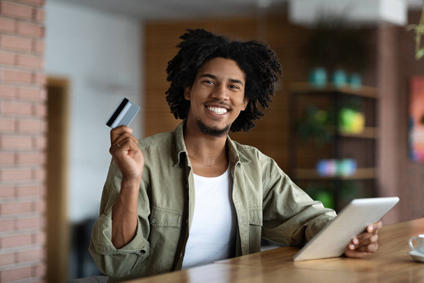 Alegre sonriente rizado milenario negro hombre celebrar tarjeta de crédito y tableta en la mesa de madera en línea de compras en la cafetería - Foto, imagen