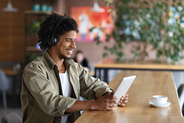 Happy smiling curly young black male in wireless headphones look at tablet and listen music in cafe - Foto, imagen