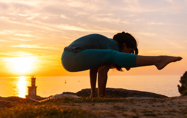 Stretching exercises, a woman doing meditation and yoga exercises on a rock at sunset next to a lighthouse in the sea, healthy and naturist life - Foto, immagini