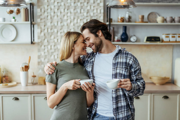 Glad european millennial wife blond and man with stubble drink coffee from cups and enjoy tender moment - Photo, Image