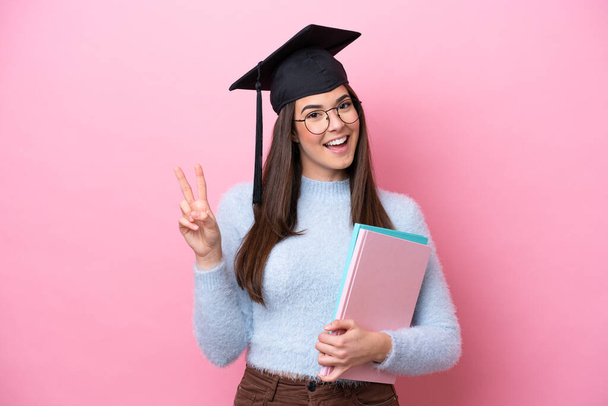 Nuori opiskelija Brasilian nainen yllään valmistui hattu eristetty vaaleanpunainen tausta hymyilee ja osoittaa voiton merkki - Valokuva, kuva