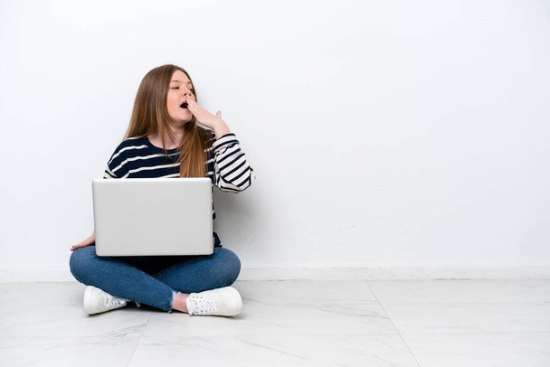 Young caucasian woman with a laptop sitting on the floor isolated on white background yawning and covering wide open mouth with hand - Fotó, kép