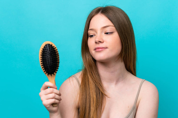 Nuori valkoihoinen nainen eristetty sinisellä taustalla hiukset kampa ja etsivät sitä - Valokuva, kuva