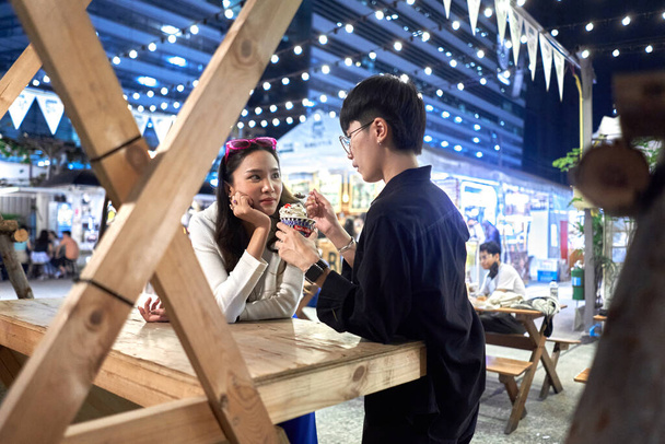 Non binary person feeding ice cream to her partner at urban night fair - 写真・画像