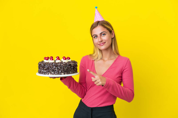 Sarışın Uruguaylı kız elinde doğum günü pastası ile sarı arka planda izole edilmiş bir şekilde bir ürün sunmak için yan tarafı işaret ediyor. - Fotoğraf, Görsel