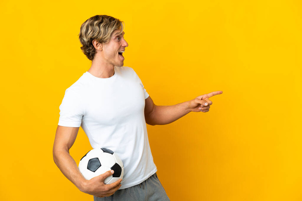 İngiliz futbolcu, izole edilmiş sarı zemin üzerinde parmak göstererek bir ürün sunuyor. - Fotoğraf, Görsel