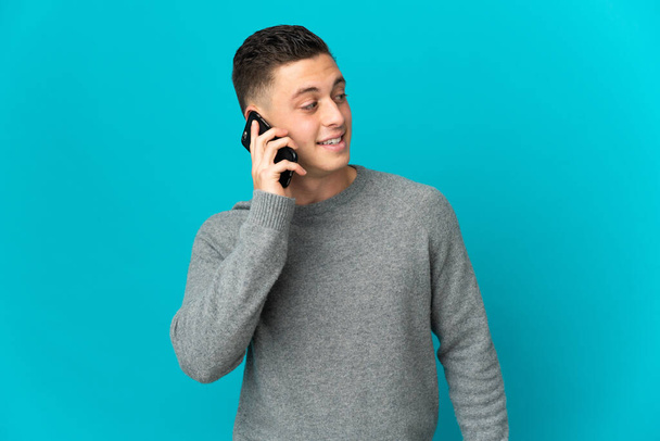 Jeune homme caucasien isolé sur fond bleu garder une conversation avec le téléphone portable avec quelqu'un - Photo, image