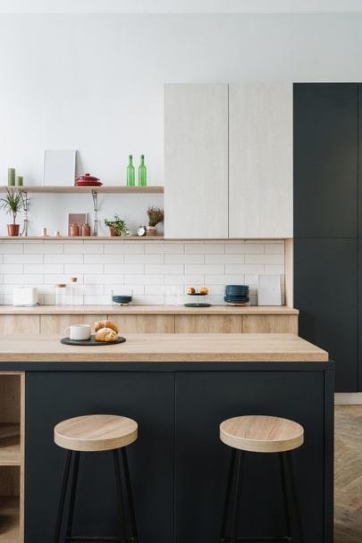Vertical shot of upscale interior with wooden furniture and bar chairs close to hardwood countertop, against shelves with kitchen utensils and home decor in cabinet. Apartment with loft design - Fotó, kép