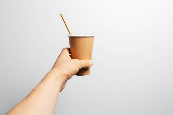 Κοντινό πλάνο του αρσενικού χεριού κρατώντας χάρτινο φλιτζάνι καφέ με καλαμάκι μπαμπού σε λευκό. - Φωτογραφία, εικόνα