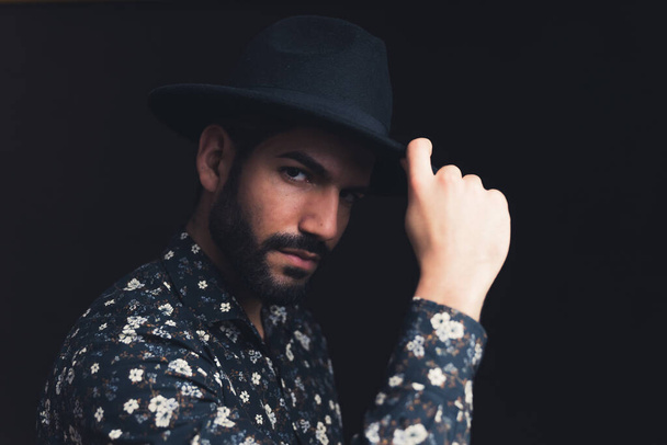 bearded Latin American handsome male person touching black hat looking severely into the camera copy space black background studio shot medium closeup - Φωτογραφία, εικόνα
