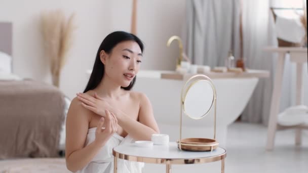 Mujer asiática joven envuelta en toalla aplicando loción de mimos en hombros y cuello, cuerpo hidratante en el baño - Imágenes, Vídeo