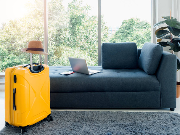 Empty dark sofa seat with passport, laptop computer, and beach hat on yellow travel suitcase on the carpet near the huge glass window at home. Ready to travel, happy holiday, summer vacation concept. - 写真・画像