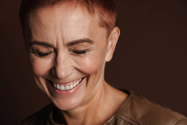 Senior ginger woman with short hair laughing while posing on camera isolated over brown wall - Fotoğraf, Görsel