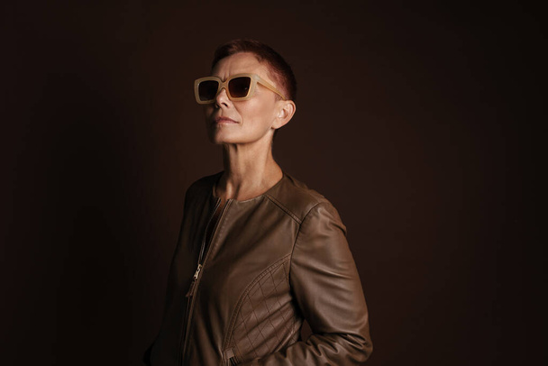Senior ginger woman in sunglasses posing and looking at camera isolated over brown wall - Fotografie, Obrázek
