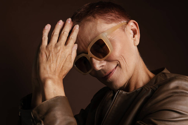 Senior ginger woman in sunglasses smiling and looking at camera isolated over brown wall - Φωτογραφία, εικόνα