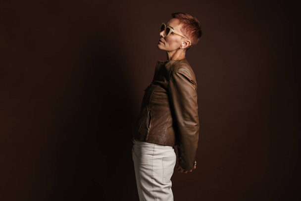 Senior ginger woman in sunglasses posing and looking aside isolated over brown wall - Fotoğraf, Görsel