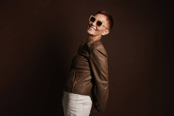 Senior ginger woman in sunglasses smiling and looking at camera isolated over brown wall - Foto, immagini