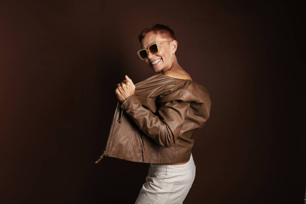 Senior ginger woman smiling while posing in leather jacket isolated over brown wall - 写真・画像