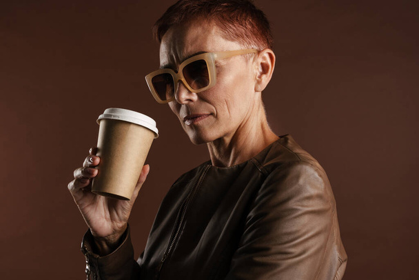 Senior ginger woman in sunglasses drinking coffee takeaway isolated over brown wall - 写真・画像