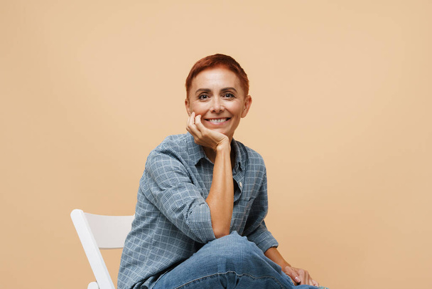 Senior ginger woman smiling at camera while sitting on chair isolated over beige wall - Foto, Bild