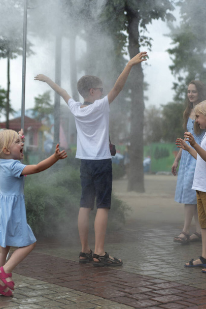 Děti se poflakují v parku a radují se z vánku. Rozstřikovač vody pro lidi v horkém dni. Procházka v parku za slunečného dne - Fotografie, Obrázek