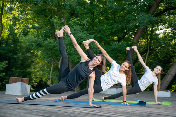 Three young woman workout in summer park doing exercise outdoor. Women does yoga asana together outside - Photo, Image