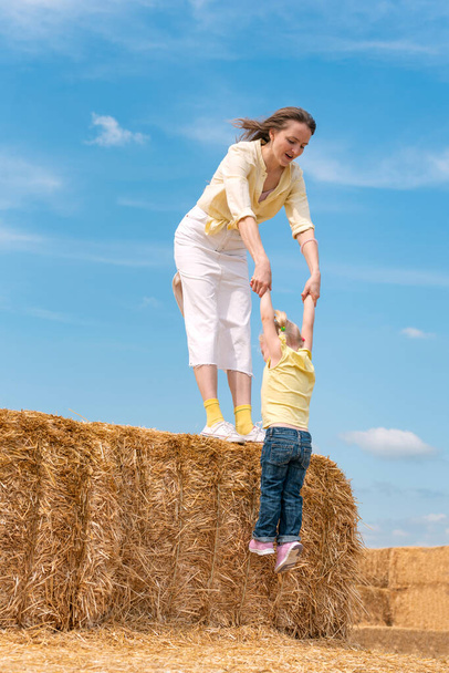 Madre joven juega con un niño pequeño en la granja en heno seco. Mamá y su hija en el pajar. Cielo azul. Día de verano - Foto, Imagen