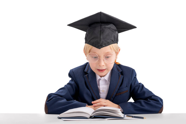 Okul üniformalı ve öğrenci şapkalı genç bir öğrenci masada oturmuş ders kitabı okuyor. Eğitim konsepti. Beyaz arkaplanda izole - Fotoğraf, Görsel