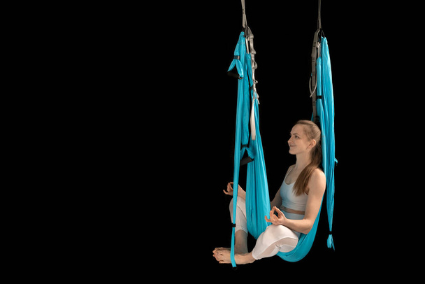Woman sits in lotus pose in hammock practicing aerial yoga black background. Copy space. Anti-gravity yoga - Photo, Image