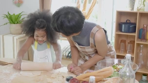 African America family with mother wearing apron rolling thresh flour for cooking with daughter together in the kitchen at home, parent and little child preparing food with fun and playful. - Footage, Video