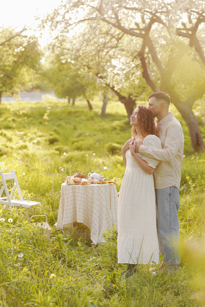 ロマンチックなデートのピクニックで美しい夏の緑の公園で食べ物や飲み物を楽しんでいる若いカップル、ハンサムな男と巻き毛の女性は屋外で抱き合っています. - 写真・画像