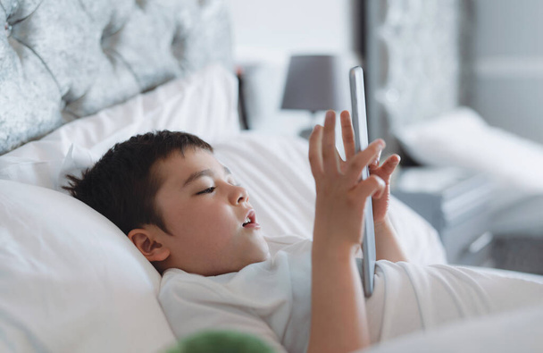 Happy Kid makaa sängyssä tilalla tabletti katsomassa sarjakuva ja jutella ystävien kanssa digitaalisessa pad, Söpö nuori poika pelaa pelejä verkossa Internetissä, Lapsi rentouttava aamulla ennen mennä kouluun - Valokuva, kuva